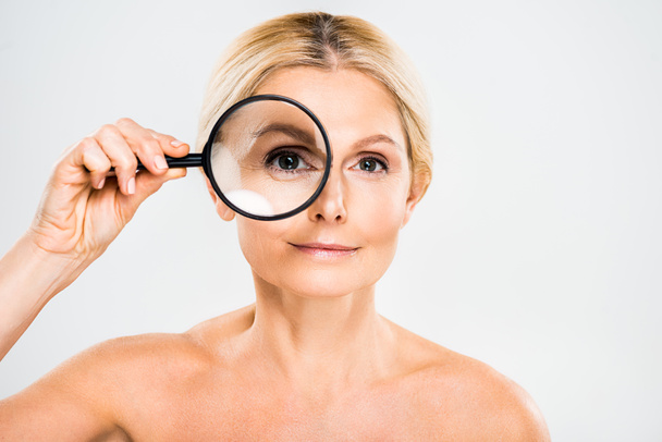 beautiful and blonde mature woman looking at camera and holding loupe on grey background  - 写真・画像