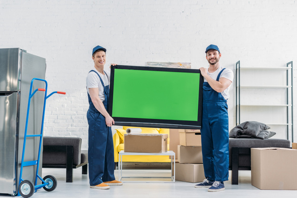 two movers in uniform looking at camera while transporting tv with green screen in apartment - Photo, Image