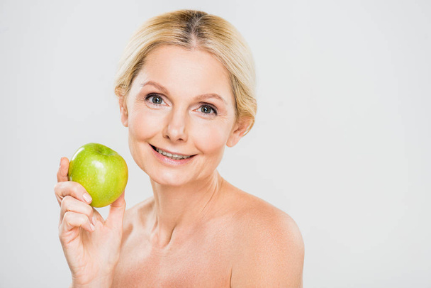beautiful and smiling mature woman holding green apple and looking at camera on grey background  - Photo, Image