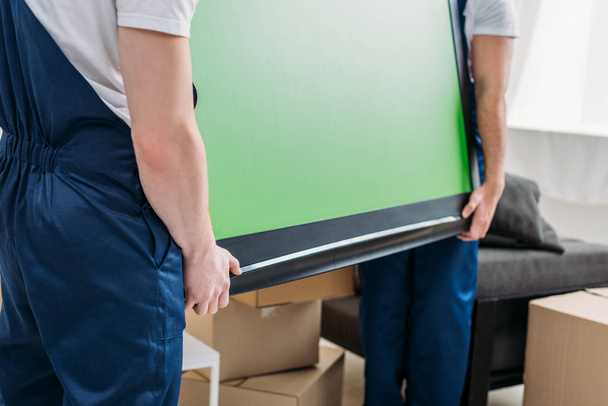 cropped view of two movers in uniform transporting tv with green screen in apartment - Foto, Imagen