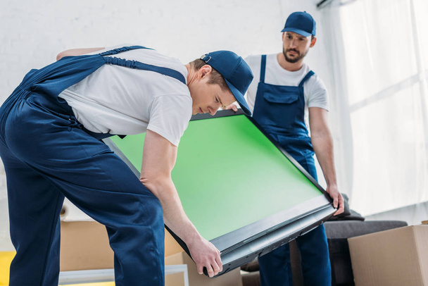 two handsome movers in uniform transporting tv with green screen in apartment - Fotoğraf, Görsel