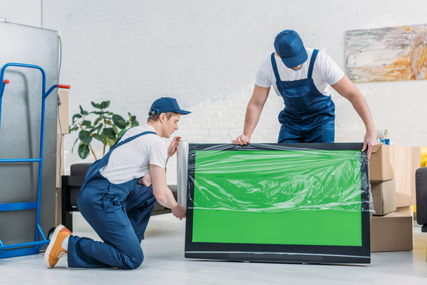 два грузчика в форме с помощью рулона стретч-пленки во время обертывания телевизор с зеленым экраном в квартире
 - Фото, изображение