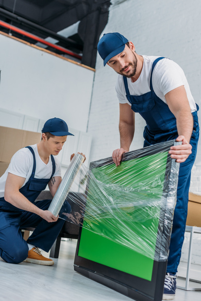 two movers using roll of stretch film while wrapping tv with green screen in apartment - Φωτογραφία, εικόνα