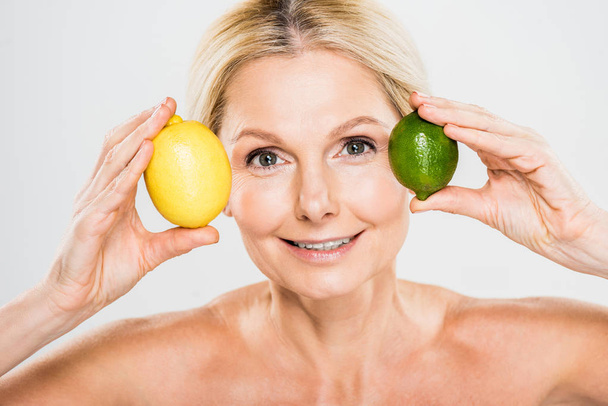 bela e sorridente mulher madura segurando verde maduro limão e limão e olhando para a câmera em fundo cinza
  - Foto, Imagem