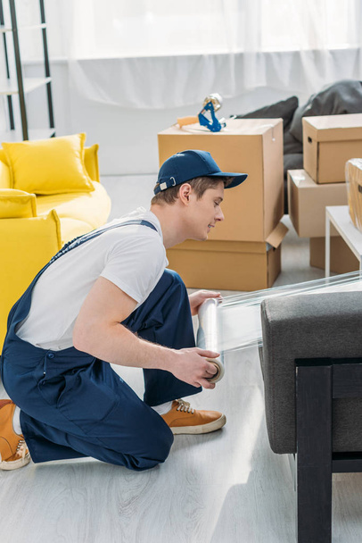 déménageur en uniforme enveloppant des meubles avec rouleau de film étirable dans l'appartement
 - Photo, image