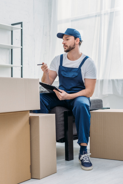 guapo mover sentado cerca de cajas de cartón y sujetando portapapeles en apartamento
 - Foto, Imagen