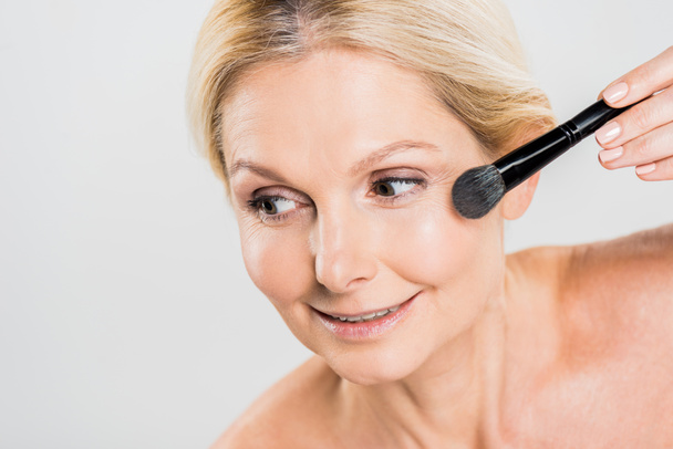 beautiful and smiling woman applying blush with cosmetic brush and looking away isolated on grey  - Foto, Imagem