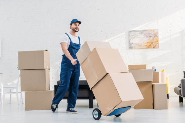 mover in uniform transporting  cardboard boxes on hand truck in apartment - Photo, Image