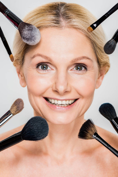 beautiful and smiling woman looking at camera with cosmetic brushes isolated on grey  - Valokuva, kuva