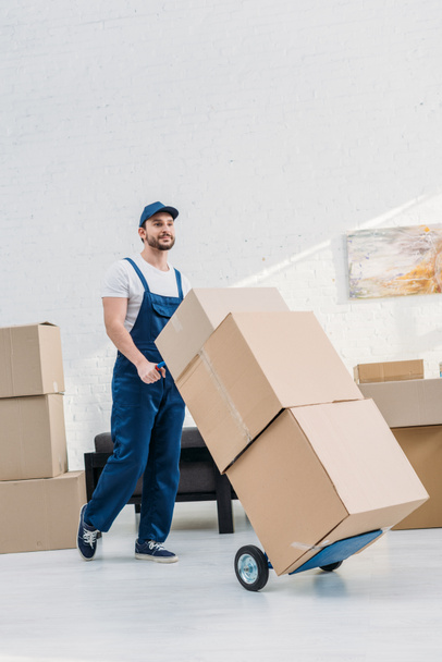 mover in uniform transporting cardboard boxes on hand truck in apartment with copy space - Photo, Image