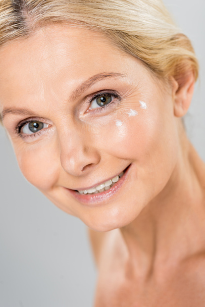 portrait of beautiful and mature woman looking at camera with cosmetic cream on face isolated on grey  - Photo, Image