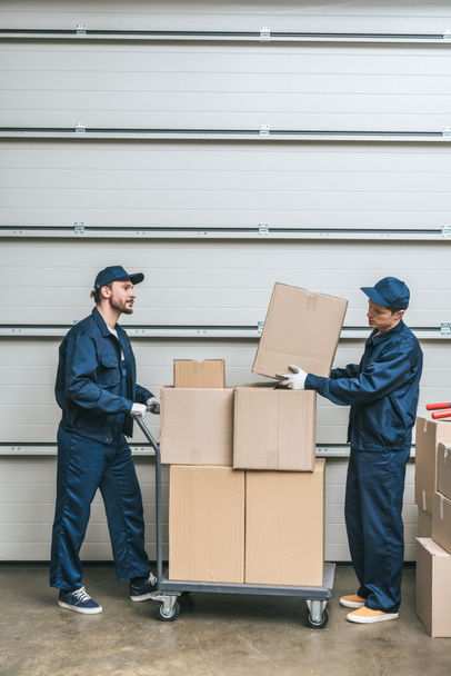 two movers in uniform transporting cardboard boxes on hand truck in warehouse with copy space - Foto, Bild