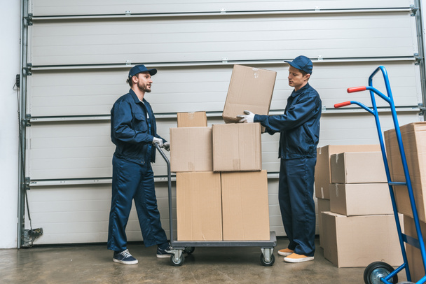 two movers in uniform transporting cardboard boxes on hand truck in warehouse with copy space - Foto, immagini