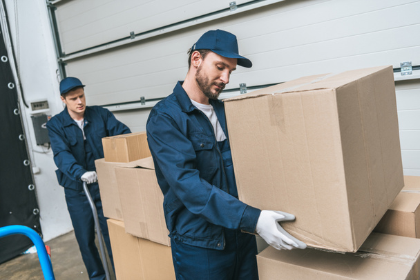 two concentrated movers in uniform transporting cardboard boxes in warehouse - Valokuva, kuva