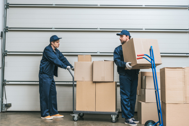 two movers in uniform looking at each other while transporting cardboard boxes with hand trucks in warehouse - Foto, immagini