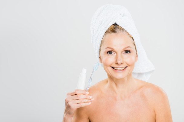 hermosa y sonriente mujer madura en toalla sosteniendo pasta de dientes y cepillo de dientes
 - Foto, Imagen