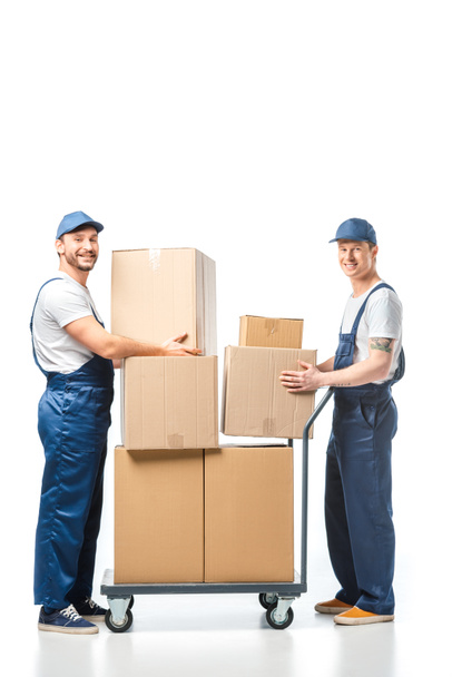 two handsome movers looking at camera while transporting cardboard boxes with hand truck on white - 写真・画像