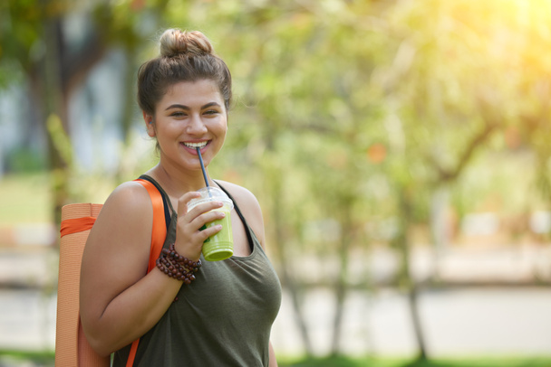Porträt einer fröhlichen jungen sportlichen Frau, die Smoothie trinkt, nachdem sie Outdoor-Yoga praktiziert hat - Foto, Bild