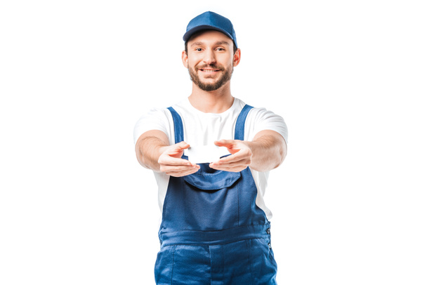 smiling handsome mover in uniform looking at camera and holding blank business card isolated on white - Φωτογραφία, εικόνα