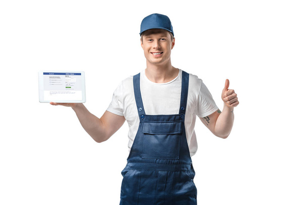 handsome smiling mover showing thumb up and presenting digital tablet with facebook app on screen isolated on white - Φωτογραφία, εικόνα