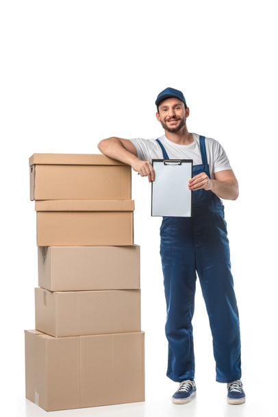 handsome mover in uniform looking at camera while holding blank clipboard near cardboard boxes isolated on white - Zdjęcie, obraz