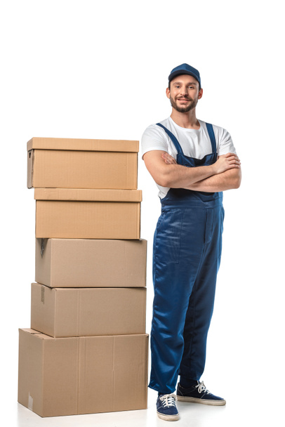 handsome mover in uniform with cardboard boxes looking at camera and smiling isolated on white - Photo, Image