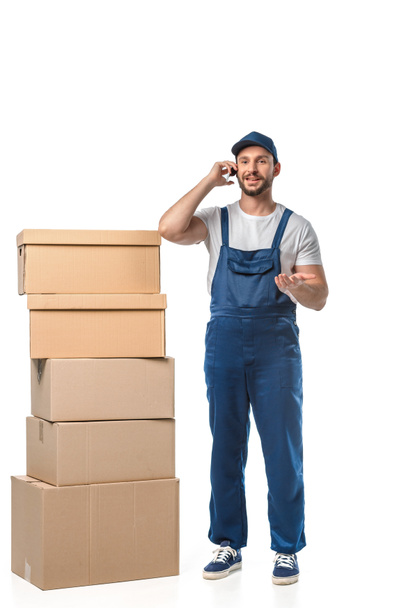 handsome smiling mover with cardboard boxes talking on smartphone and gesturing with hand isolated on white - 写真・画像