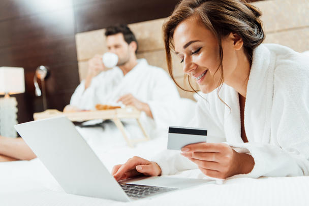 επιλεκτική εστίαση του ευτυχισμένη γυναίκα που χρησιμοποιούν φορητό υπολογιστή ενώ κρατάτε πιστωτική κάρτα κοντά σε φίλο  - Φωτογραφία, εικόνα