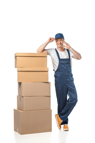 guapo sonriente mover en uniforme con cajas de cartón hablando en teléfono inteligente aislado en blanco
 - Foto, Imagen