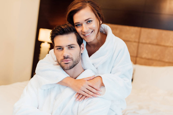 happy girlfriend in white bathrobe hugging handsome boyfriend and looking at camera  - Photo, Image