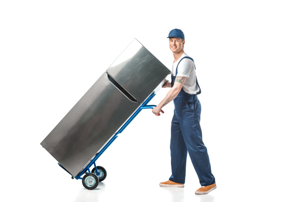 handsome mover in uniform looking at camera and transporting refrigerator on hand truck isolated on white - Фото, изображение