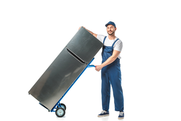 handsome mover in uniform looking at camera and transporting refrigerator on hand truck isolated on white - 写真・画像