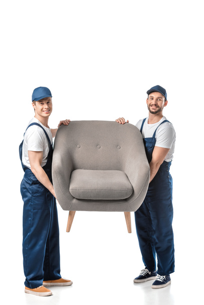 two smiling movers looking at camera and transporting grey armchair isolated on white - 写真・画像