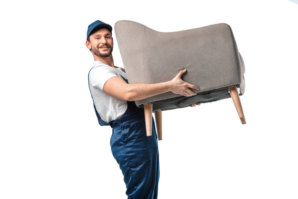 handsome mover in uniform looking at camera while carrying grey armchair isolated on white - Photo, Image