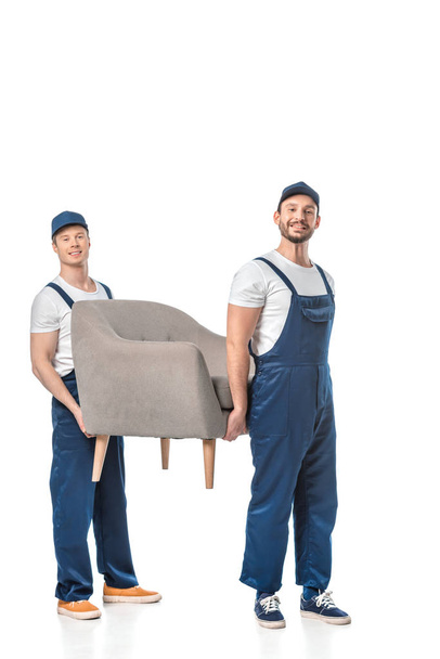 two smiling movers looking at camera and transporting grey armchair isolated on white - Photo, Image