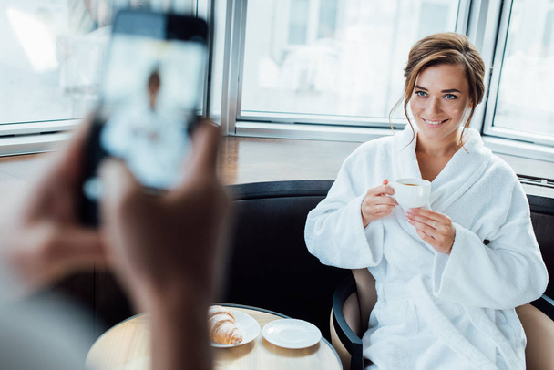 cropped view of man taking photo of attractive girlfriend holding cup while sitting in bathrobe in hotel  - Foto, Imagem