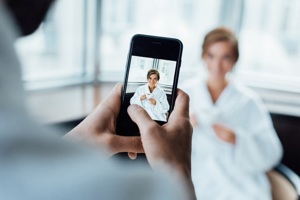 selective focus of man taking photo of attractive girlfriend holding cup while sitting in bathrobe in hotel  - Photo, image