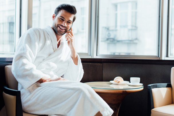 cheerful bearded man in white bathrobe sitting on chair and talking on smartphone  - Valokuva, kuva