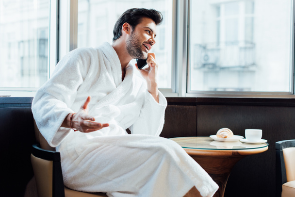 szczęśliwy Brodaty mężczyzna w biały szlafrok rozmowy na smartfon w pobliżu stolik ze śniadaniem - Zdjęcie, obraz