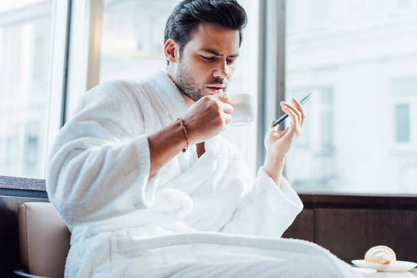 hezký vousatý muž v bílém županu držení smartphone a šálku s kávou - Fotografie, Obrázek