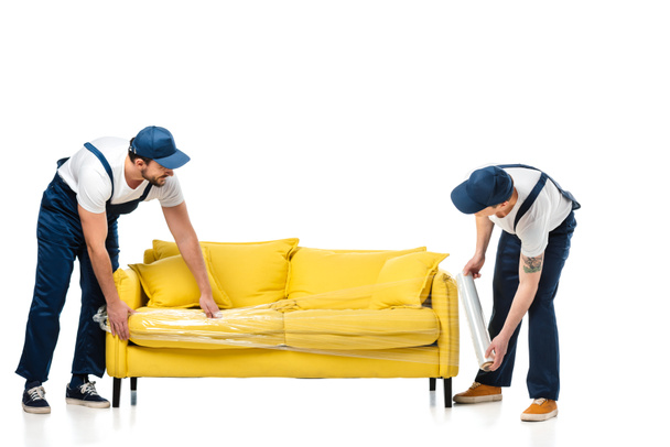 два грузчика обертывают желтый диван с рулоном стретч-пленки на белом
 - Фото, изображение