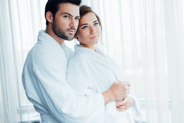 handsome man hugging happy woman in white bathrobe in hotel  - Zdjęcie, obraz