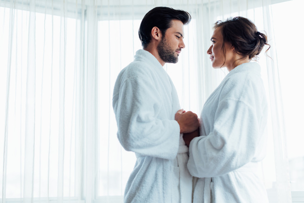 handsome bearded man holding hands with happy woman in white bathrobe in hotel  - Foto, afbeelding
