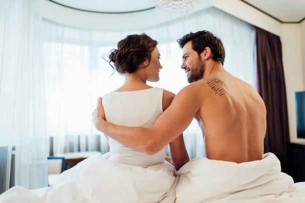 tattooed boyfriend hugging attractive brunette woman while sitting on bed  - Fotó, kép