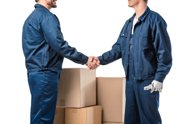 cropped view of two movers in uniform shaking hands near cardboard boxes isolated on white - Photo, Image