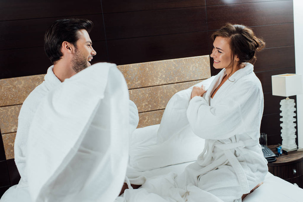 χαρούμενο ζευγάρι στο μπουρνούζια έχοντας μαξιλάρι καταπολέμηση σε κρεβάτι στο ξενοδοχείο  - Φωτογραφία, εικόνα