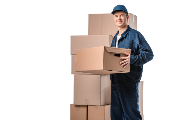 handsome mover in uniform carrying cardboard box and looking at camera isolated on white with copy space - Photo, Image