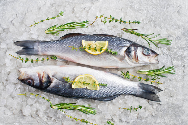 Raw sea bass on ice with rosemary, thyme and lemon. Gray background, top view. - Photo, image