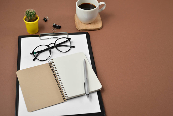 Mockup workspace with blank clip board, office supplies, cactus, pen, coffee cup, notepad and eyeglasses on brown background. Flat lay, top view, copy space - Photo, Image