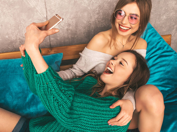 Two young beautiful smiling gorgeous girls in trendy summer hipster clothes.  Sexy carefree women posing in interior and taking selfie. Positive models having fun with smartphone - Fotó, kép